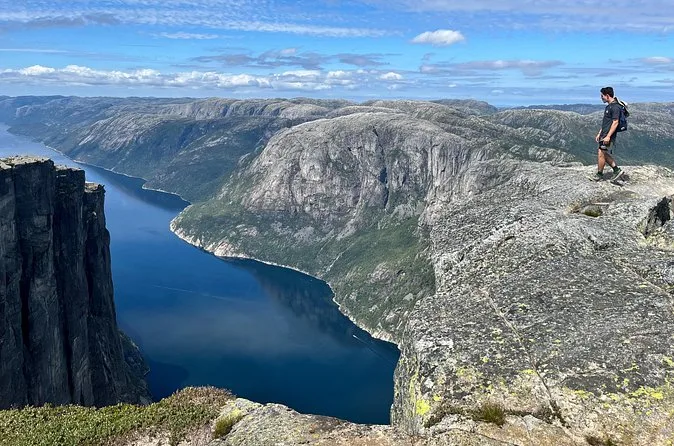 A hiker standing on a granite ledge above a long blue fjord between steep mountain walls