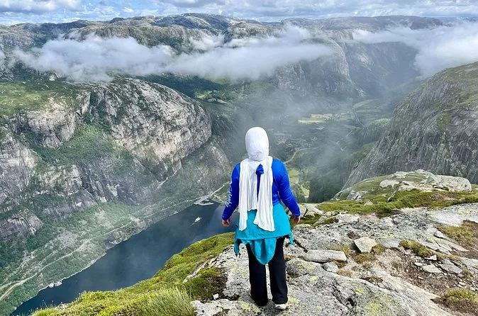 A hiker in a white headscarf standing on a rock ledge looking at cloud drifting over a fjord valley