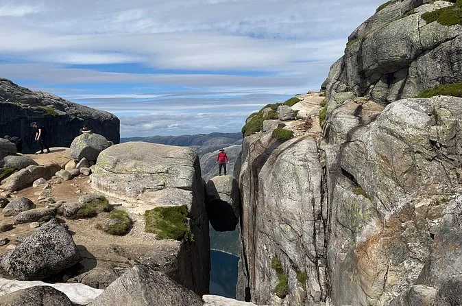 Der Kjeragbolten in einem tiefen Felsspalt, von der Seite gesehen, dahinter ein kleiner Wanderer auf den Felsen