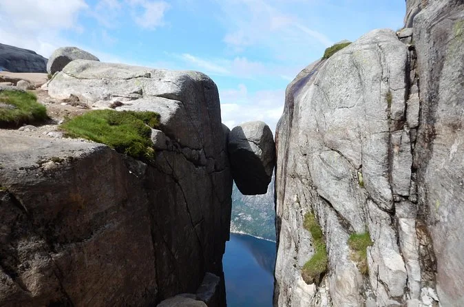 The boulder wedged between two rock walls, with the fjord visible far below through the gap