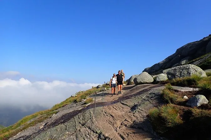Zwei Wanderer gehen in der Sonne einen breiten Granitrücken hinauf, unten füllen Wolken das Tal