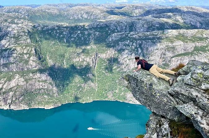 Ein Wanderer liegt auf einem Felsvorsprung über einem türkisfarbenen Fjord, tief unten ein kleines Boot
