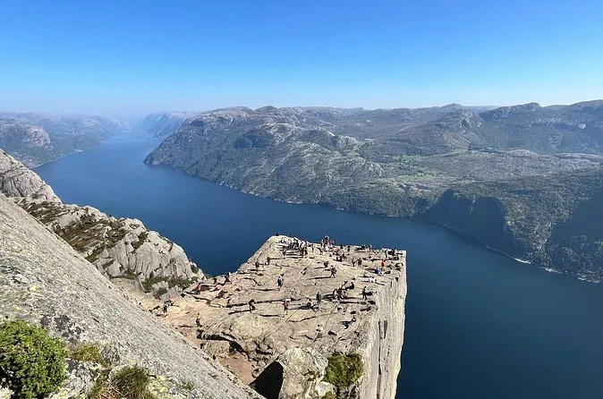 Der Preikestolen von einem höheren Felsen aus gesehen, eine flache Plattform mit Menschen über dem tiefblauen Fjord
